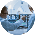 In Control Room Doctor and Radiologist Discuss Diagnosis while Watching Procedure and Monitors Showing Brain Scans Results, In the Background Patient Undergoes MRI or CT Scan Procedure 