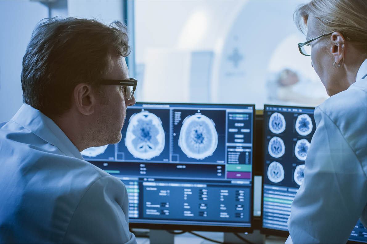 In Control Room Doctor and Radiologist Discuss Diagnosis while Watching Procedure and Monitors Showing Brain Scans Results, In the Background Patient Undergoes MRI or CT Scan Procedure.