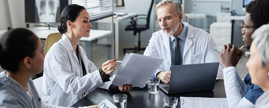 Young female clinician showing medical documents to experienced male doctor sitting by table in front of group of intercultural colleagues