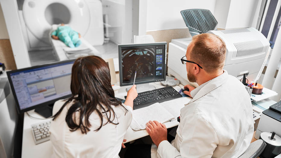 Back view of two doctors sitting, working at computer, examining MRI results. Specialists wearing white robes, consulting, woman pointing by pen, patient lying. Concept of medicine.