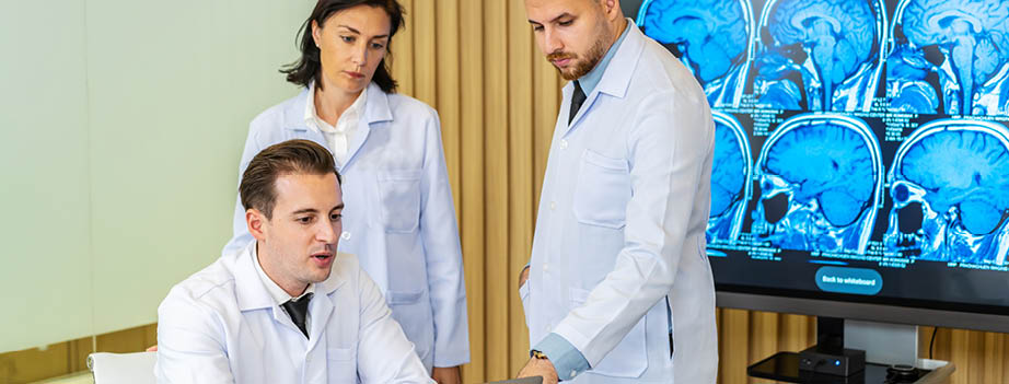 Team of three doctors discusses the results of an MRI scan of the patient's head in the consulting room of modern hospital, physician discussing about treatment for patient looking in laptop