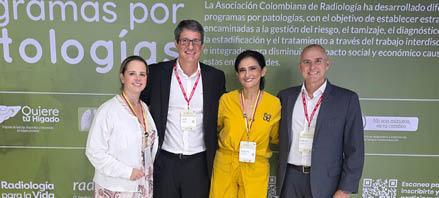 Quatro adultos com crach s posam sorrindo em frente a um painel verde com texto sobre programas para patologias da Asociaci n Colombiana de Radiolog a. Descri  o gerada por IA