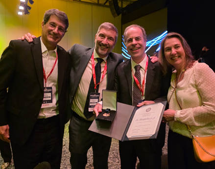 Quatro adultos sorridentes est o juntos em um ambiente interno, dois deles seguram um certificado e uma medalha, todos usam crach s vermelhos pendurados no pesco o. Descri  o gerada por IA