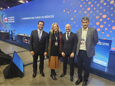 Quatro adultos vestidos formalmente est o em p sorrindo em um evento com um grande painel azul ao fundo que exibe texto relacionado a um congresso m dico.  Descri  o gerada por IA