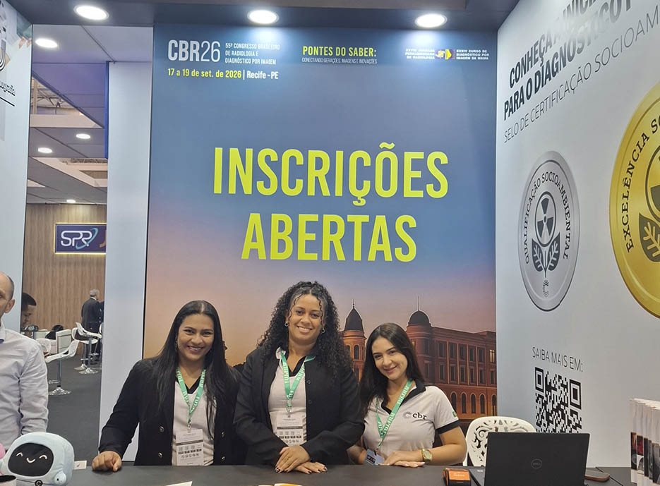 Tr s pessoas com crach s est o sentadas em uma mesa em frente a um painel azul com texto amarelo que anuncia inscri es abertas para um evento em Recife.  Descri  o gerada por IA